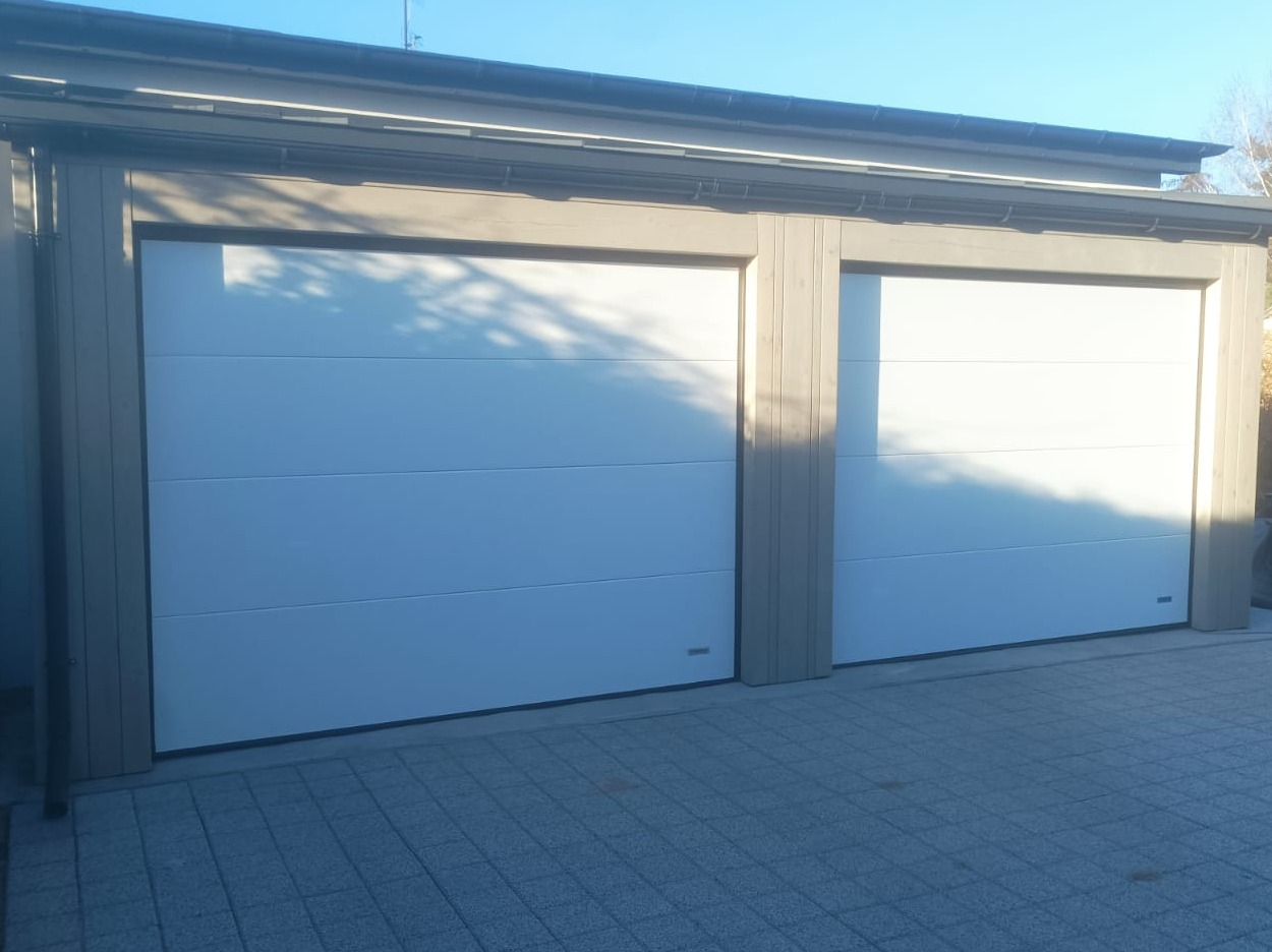 Dwie białe segmentowe bramy garażowe z szarymi obramowaniami, widoczne z zewnątrz na tle kostki brukowej i cienia drzew.