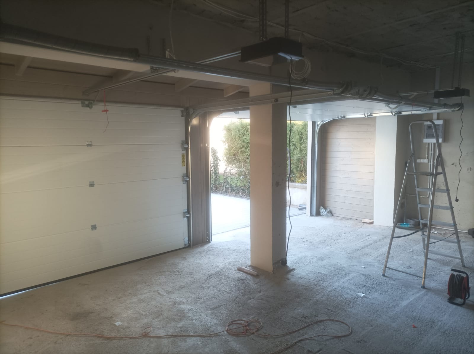 Wnętrze garażu w trakcie montażu białej segmentowej bramy, widoczne elementy konstrukcyjne pod sufitem, drabina oparta o ścianę, surowy beton na podłodze i częściowo otwarte wrota z widokiem...