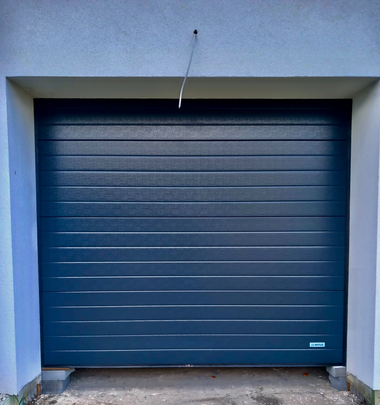 Zamknięta, segmentowa brama garażowa w kolorze grafitowym, z poziomymi przetłoczeniami. Widoczny fragment elewacji i betonowe podłoże.