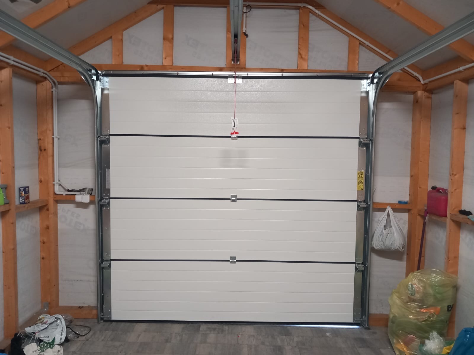 Zamknięta, segmentowa brama garażowa w jasnym garażu z drewnianą konstrukcją. Widoczne prowadnice i elementy montażowe. U dołu śmieci i plastikowy kanister.