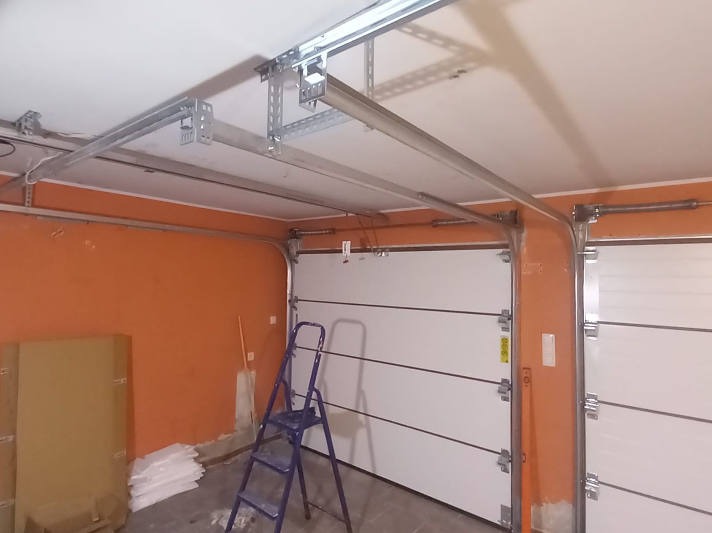 Montaż bramy garażowej segmentowej: widoczne szyny i elementy mocujące do sufitu, częściowo zamontowane panele bramy. W tle pomarańczowa ściana i drabina.