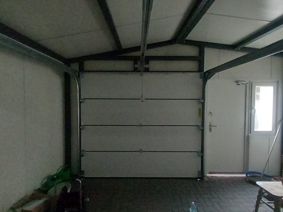 Zamknięta segmentowa brama garażowa w Poznań. Widoczne prowadnice, konstrukcja dachu i drzwi wejściowe z oknem. Brama w kolorze białym.