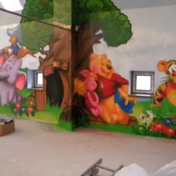 Malowidło ścienne przedstawiające postacie z Kubusia Puchatka: Prosiaczka, Puchatka, Tygryska i słonia z kapeluszem, wykonane w pokoju dziecięcym; widoczne okna i karton na podłodze.