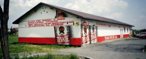 Budynek z dużym, wielkoformatowym graffiti reklamującym piwo Tyskie, z widocznymi napisami i logo marki na białym tle z czerwonym pasem na dole.