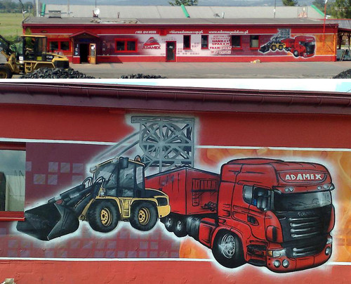 Budynek z muralem przedstawiającym ciężarówkę, ładowarkę i konstrukcję kopalnianą, wykonanym w technice graffiti. Na budynku widoczna nazwa firmy 'ADAMEX' i dane kontaktowe.