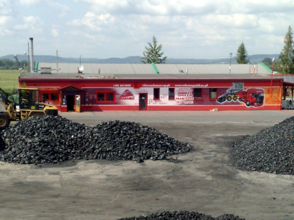 Budynek firmy z reklamą malarską na elewacji przedstawiającą lawetę i koparkę, przed budynkiem hałdy węgla.