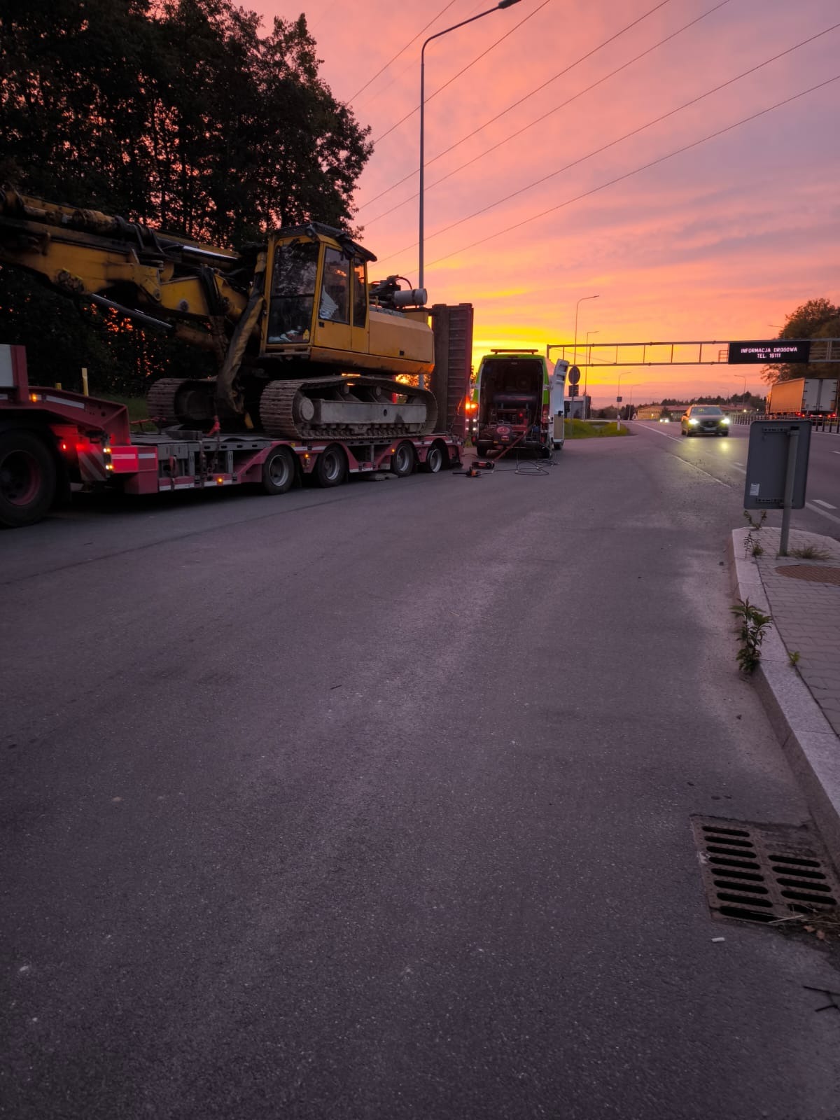 Żółta koparka gąsienicowa przewożona na czerwonej niskopodwoziowej lawecie na tle zachodzącego słońca, obok zaparkowany van z narzędziami.