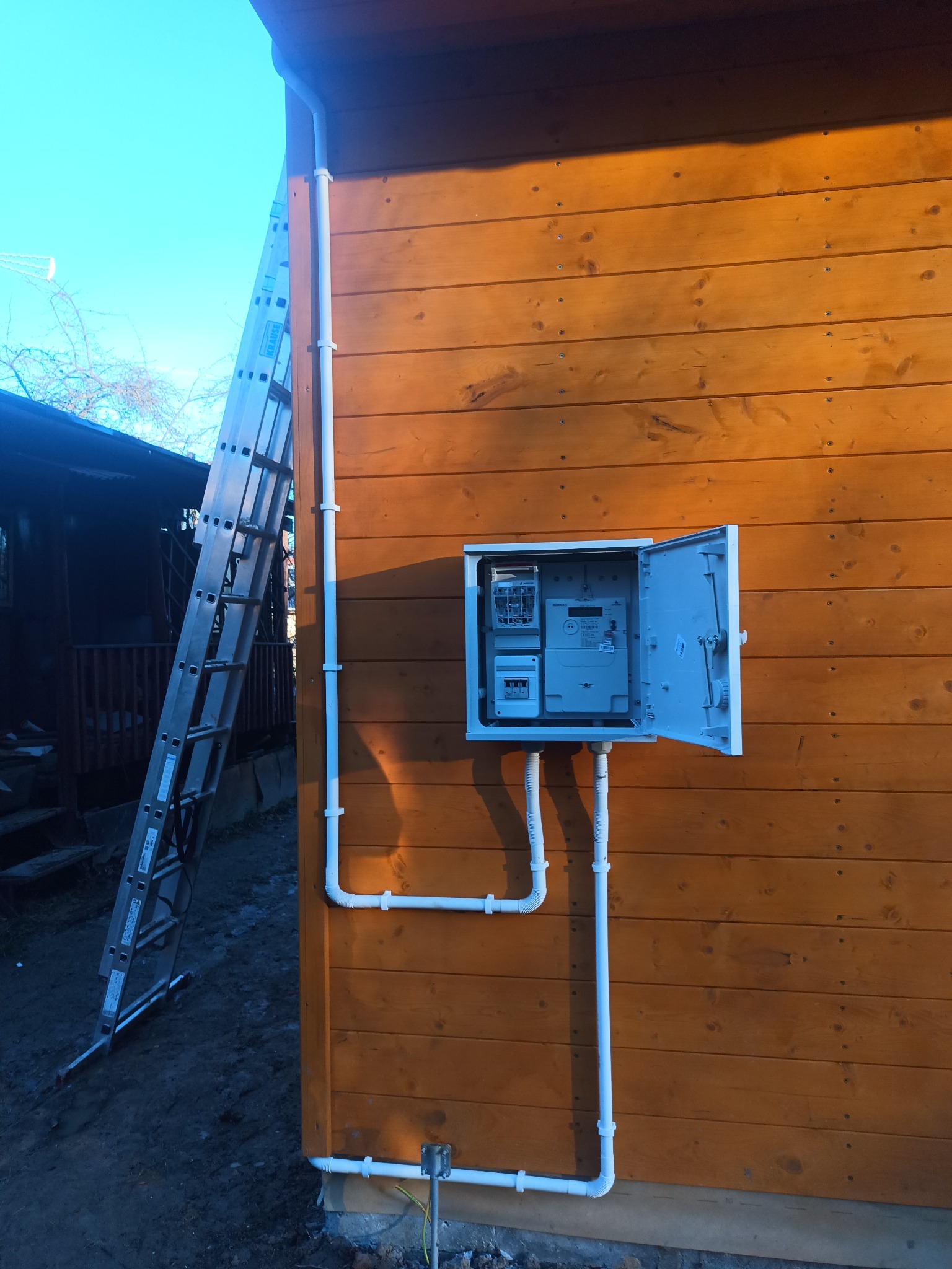 Zewnętrzna skrzynka elektryczna z otwartymi drzwiczkami na drewnianej elewacji. Widoczne białe rury PCV. Obok drabina oparta o ścianę. Instalacja wykonana na elewacji budynku.