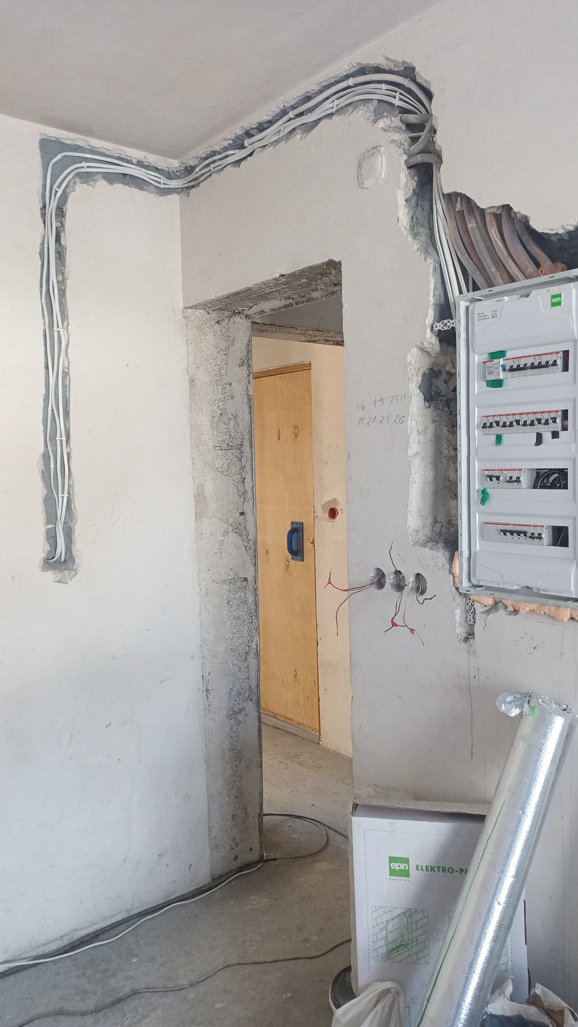 Białe przewody elektryczne ułożone w bruzdach na ścianie, z zamontowaną skrzynką rozdzielczą w surowym stanie budynku przed wykończeniem.
