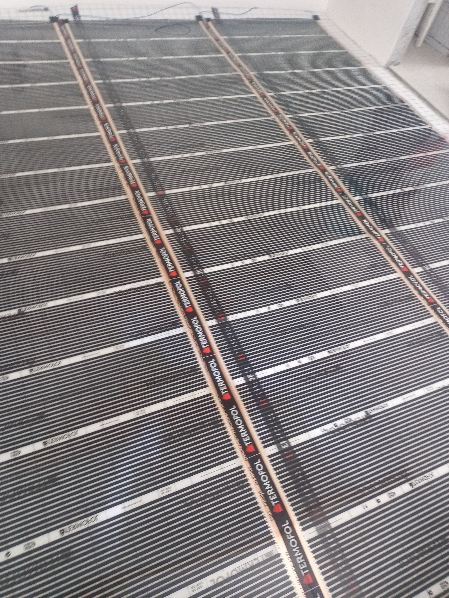 Rozłożona mata grzewcza Termofol na podłodze, przygotowana do montażu. Widoczne czarne pasy grzewcze i brązowe listwy mocujące.