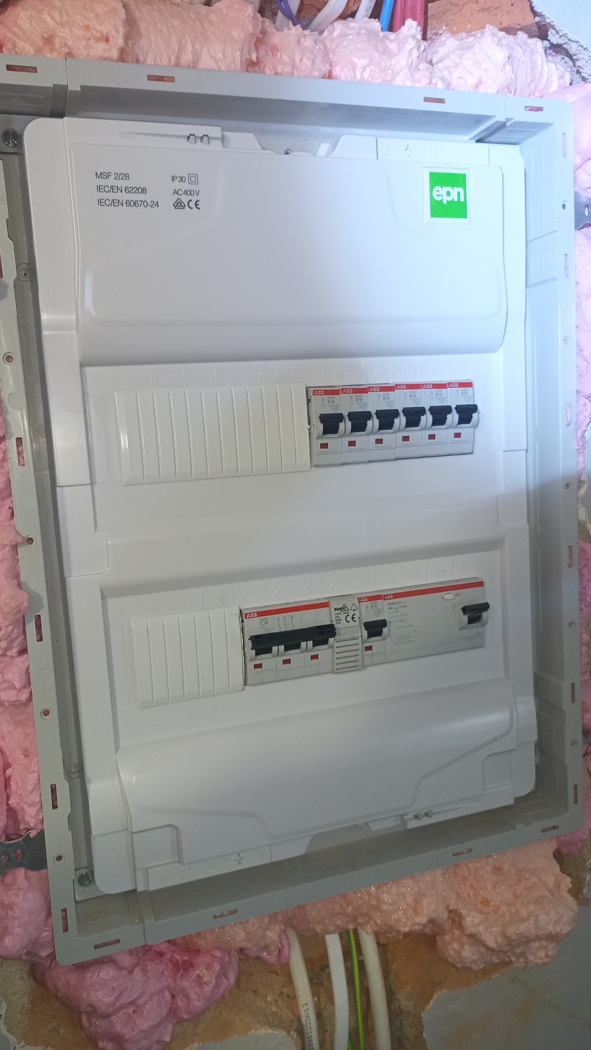 Zamontowana, biała rozdzielnica elektryczna EPN z bezpiecznikami ABB, osadzona w ścianie z pianką montażową, widoczne kable. Instalacja w trakcie prac wykończeniowych.