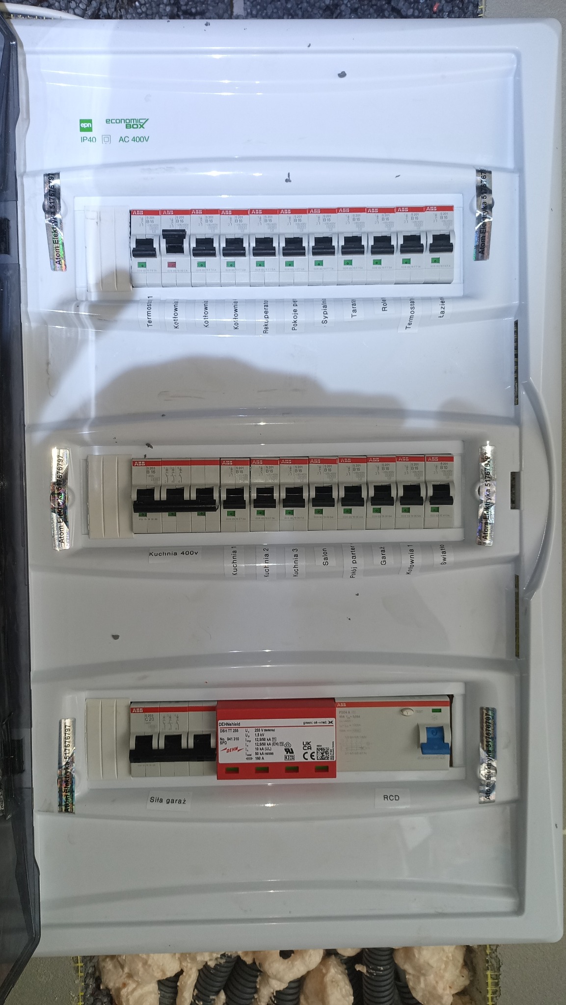 Rozdzielnica elektryczna z bezpiecznikami ABB, opisana dla różnych obwodów (kuchnia, salon, garaż), z zabezpieczeniem przeciwprzepięciowym Dehn. Widoczne kable w pianie.