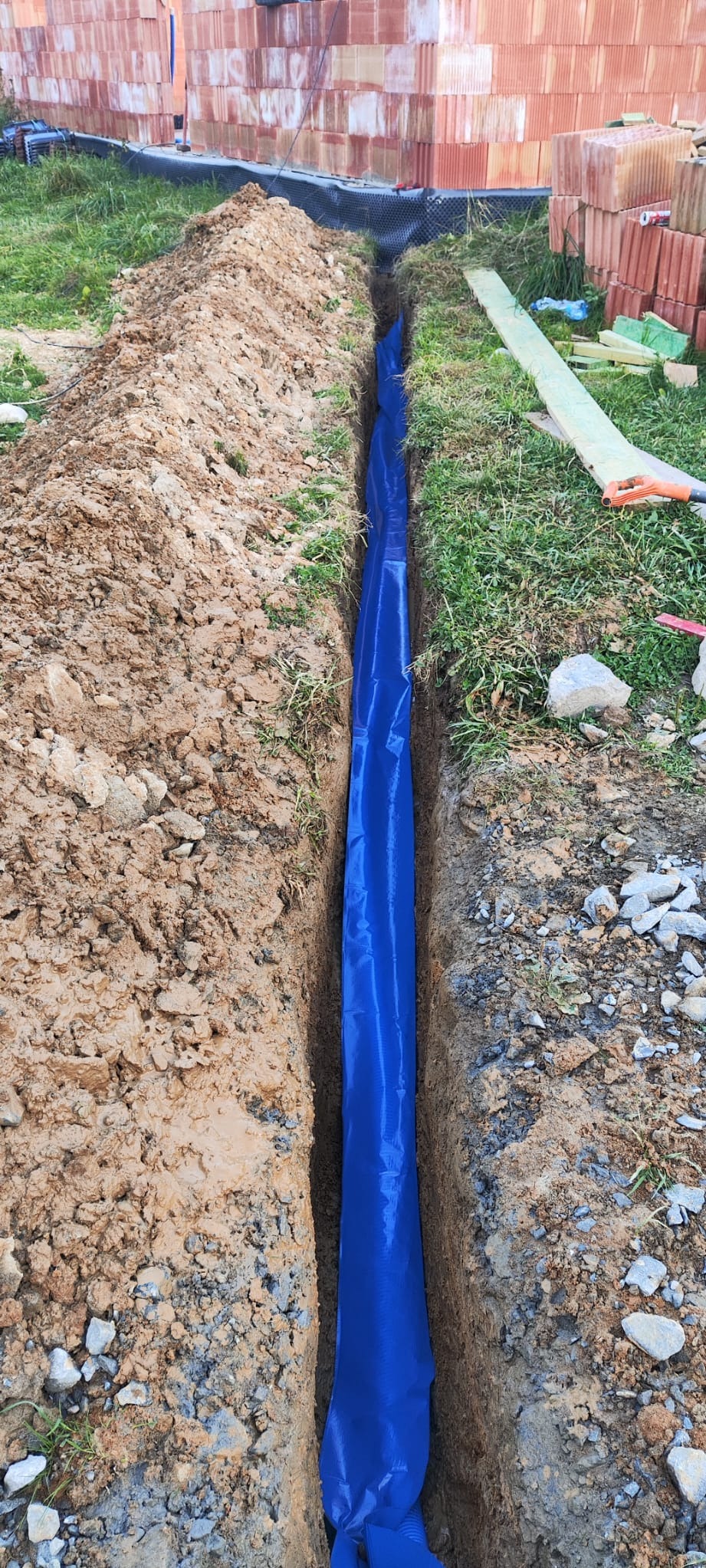 Wykop z niebieską folią ochronną na dnie, prawdopodobnie pod instalację elektryczną. Widoczne cegły i trawa w tle.