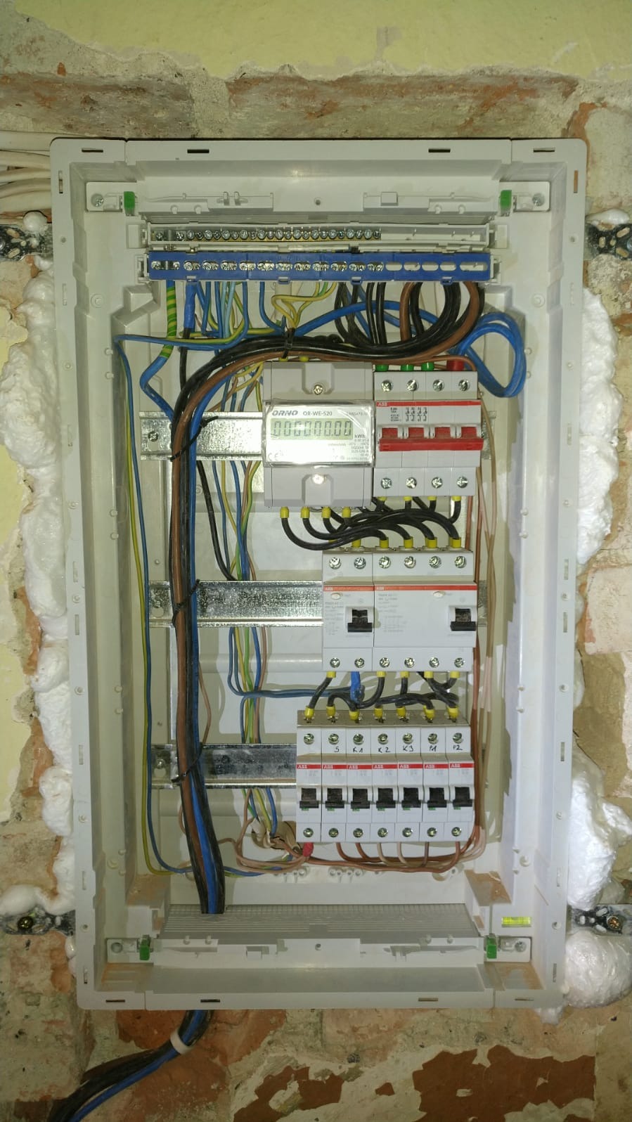Rozdzielnica elektryczna ORNO z licznikiem i zabezpieczeniami ABB, zamontowana na ścianie z cegły, uszczelniona pianką montażową. Widoczne przewody w różnych kolorach.