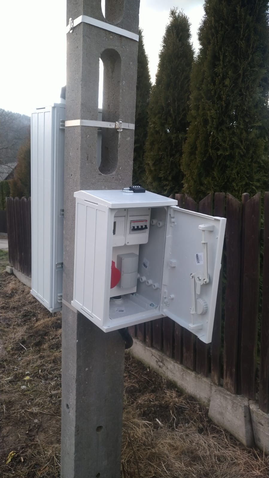 Szara betonowa słup z zamontowaną białą skrzynką elektryczną, otwartą, ukazującą liczniki i bezpieczniki. Widoczne drzewa w tle.
