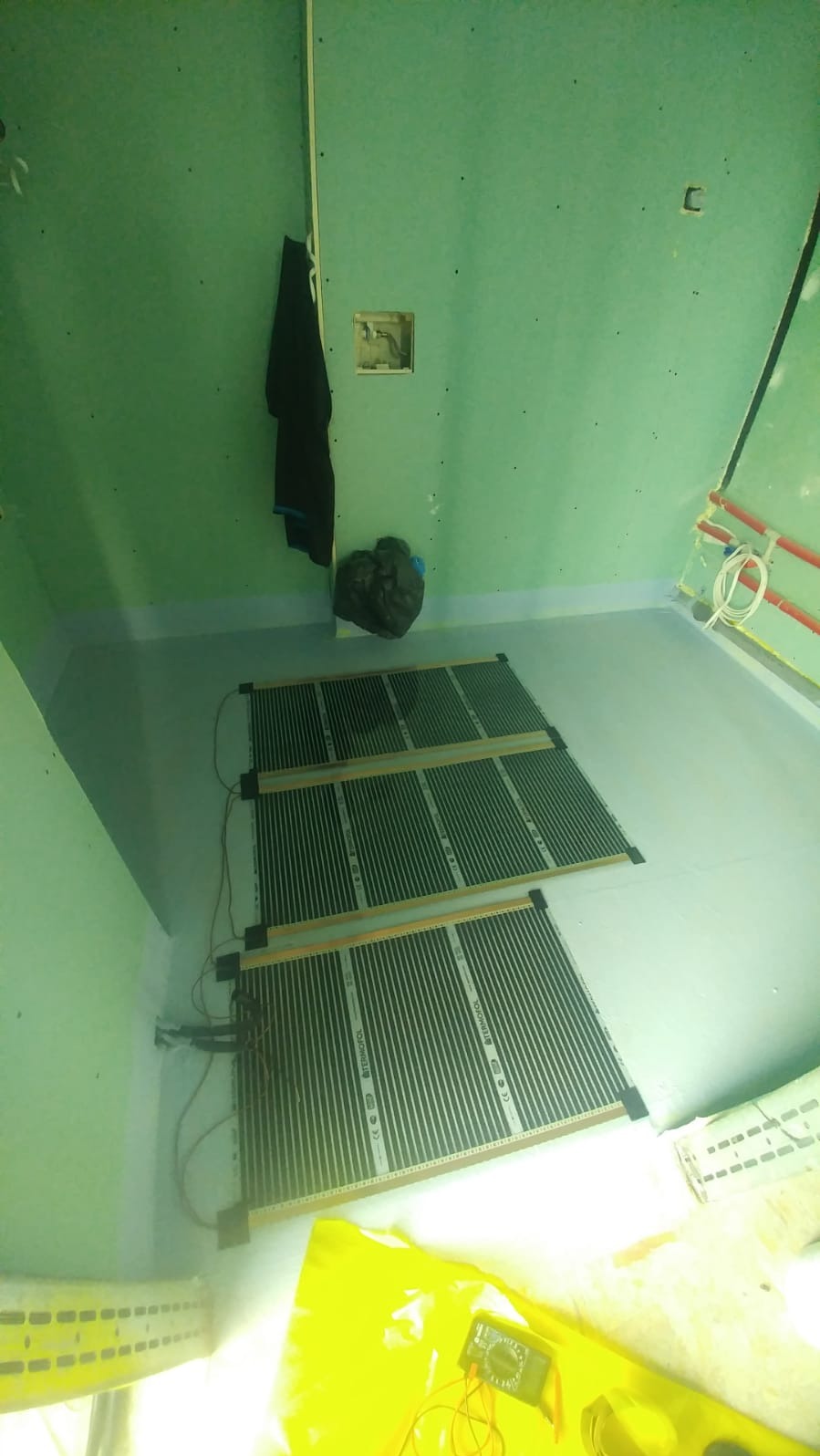 Montaż elektrycznego ogrzewania podłogowego w łazience. Widoczne maty grzewcze na podłodze, ściany w zielonych płytach kartonowo-gipsowych, miernik elektryczny w rogu.
