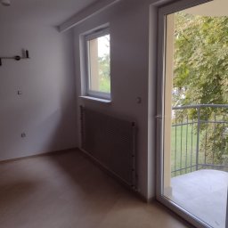 ŁUKASZ STAWSKI - Pusty pokój z jasną drewnianą podłogą, grzejnikiem pod oknem i wyjściem na balkon z widokiem na drzewa.