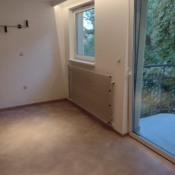 ŁUKASZ STAWSKI - Pusty pokój po remoncie z nową podłogą imitującą drewno, białymi ścianami, grzejnikiem i dużym oknem balkonowym z widokiem na zieleń.