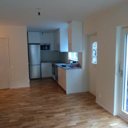 ŁUKASZ STAWSKI - Nowoczesna, jasna kuchnia z białymi szafkami i parkietem ułożonym w jodełkę. Widoczny fragment salonu z drzwiami balkonowymi. Brak wykończenia sufitu w postaci lampy.