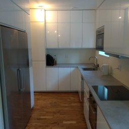 ŁUKASZ STAWSKI - Nowoczesna kuchnia z białymi, błyszczącymi szafkami, drewnianą podłogą parkietową i stalową lodówką, oświetlona centralnym światłem, z widocznymi detalami jak bateria kuchenna, czajnik elektryczny...