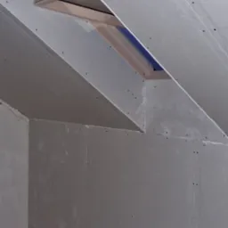 Montaż płyt gipsowo-kartonowych na poddaszu z oknem dachowym, widoczne łączenia płyt i ślady szpachlowania, przygotowanie do dalszych prac wykończeniowych.