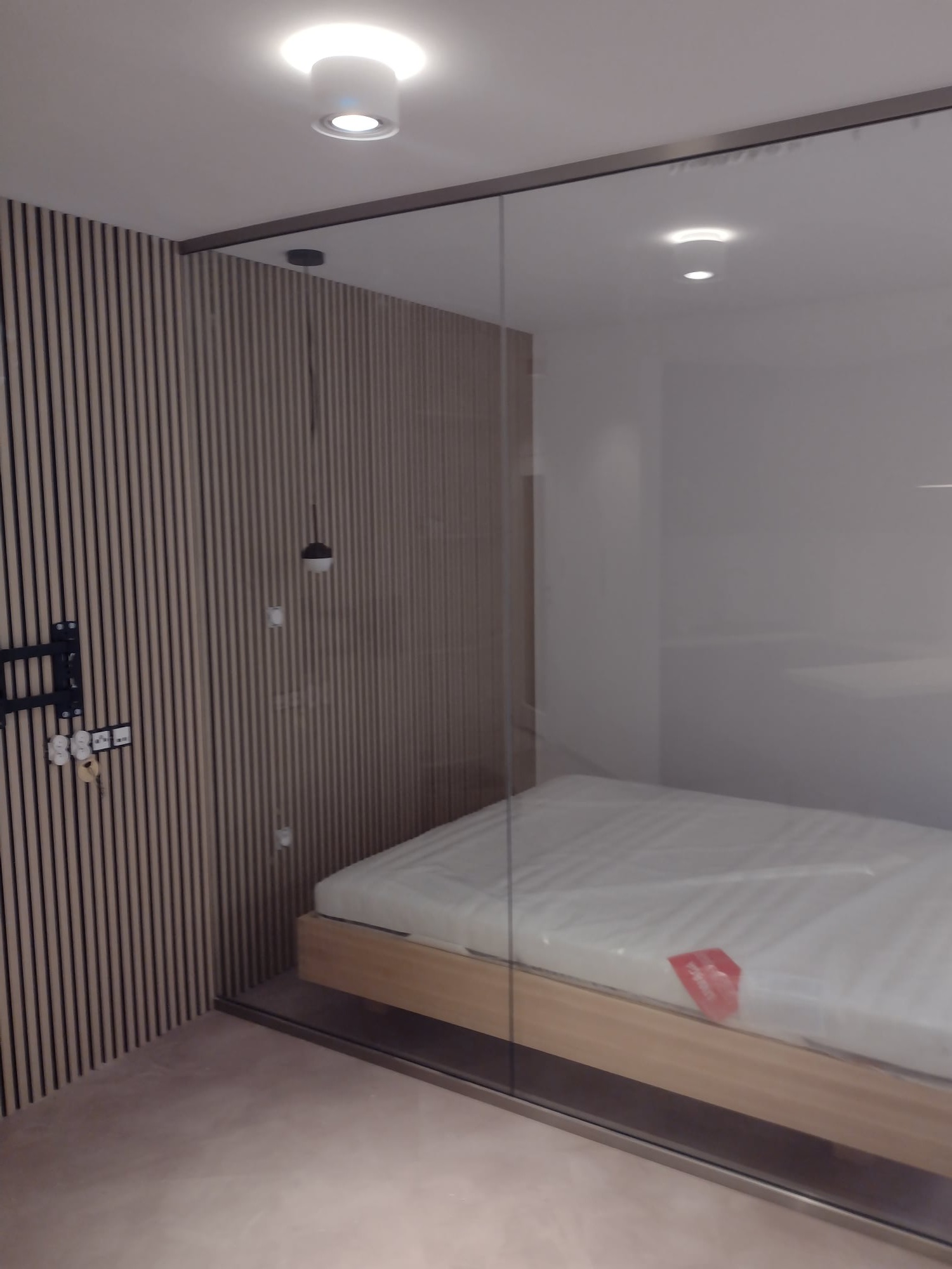 Nowoczesna sypialnia z drewnianą ścianą z pionowych paneli, szklaną ścianą działową i minimalistycznym oświetleniem sufitowym.