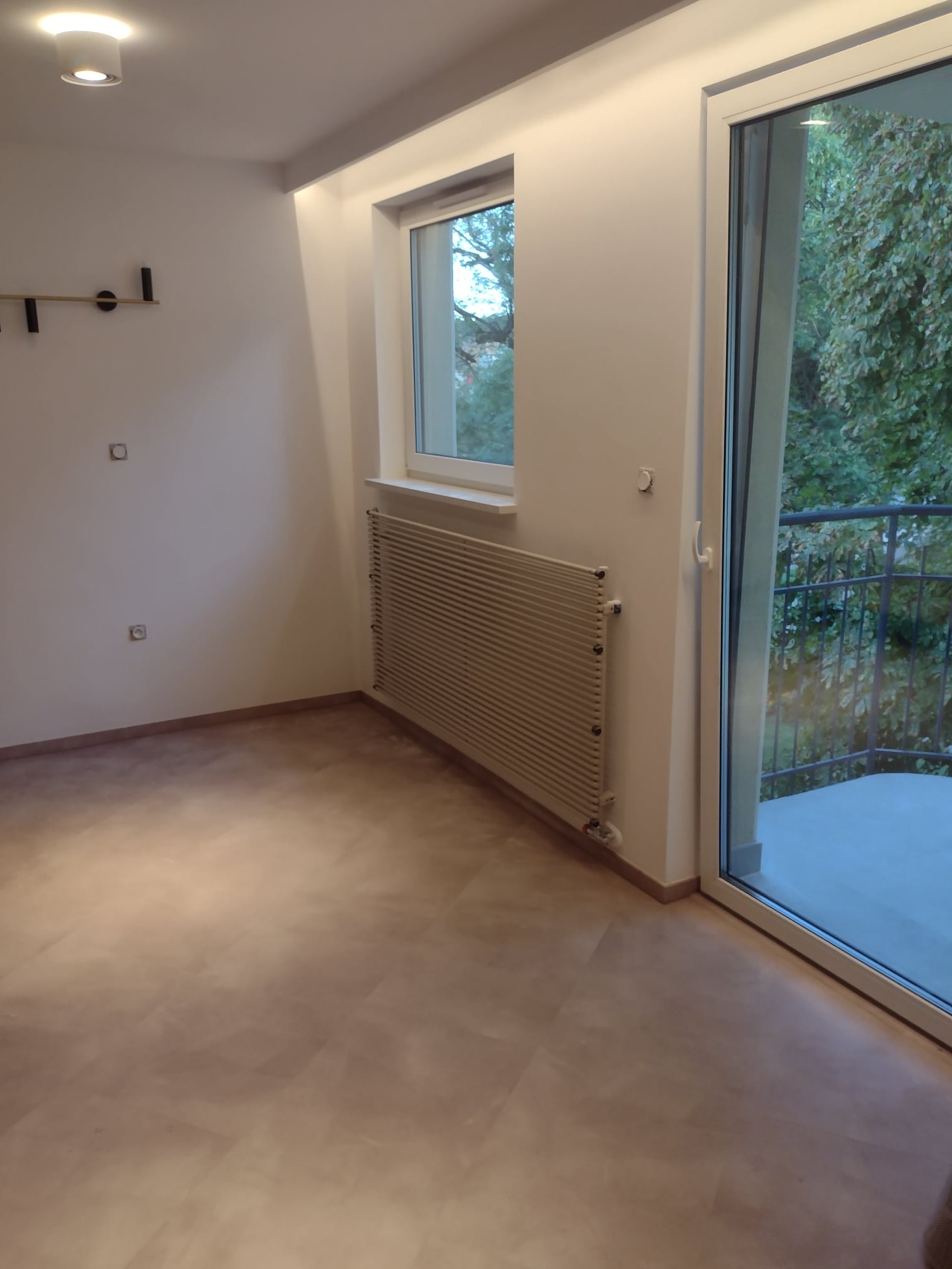 Pusty pokój po remoncie z nową podłogą imitującą drewno, białymi ścianami, grzejnikiem i dużym oknem balkonowym z widokiem na zieleń.