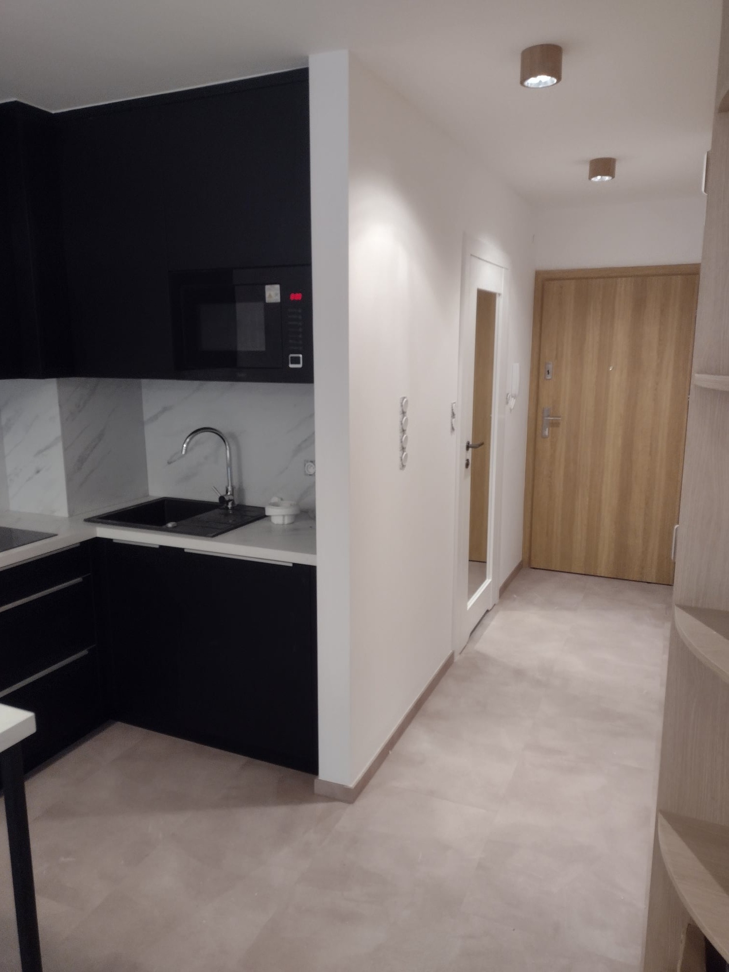 Nowoczesna kuchnia z czarnymi szafkami i marmurowym backsplashiem, widok na przedpokój z drewnianymi drzwiami i oświetleniem punktowym.