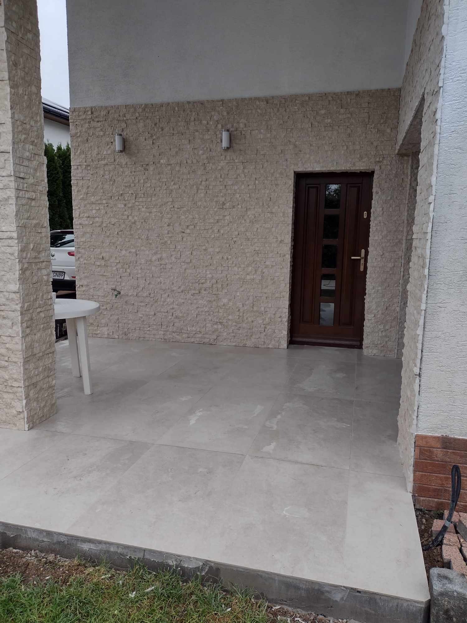 Wykończone wejście do domu z kamienną elewacją, drewnianymi drzwiami i jasną podłogą z płytek. Widoczny fragment białego stolika i kolumna z kamienia.