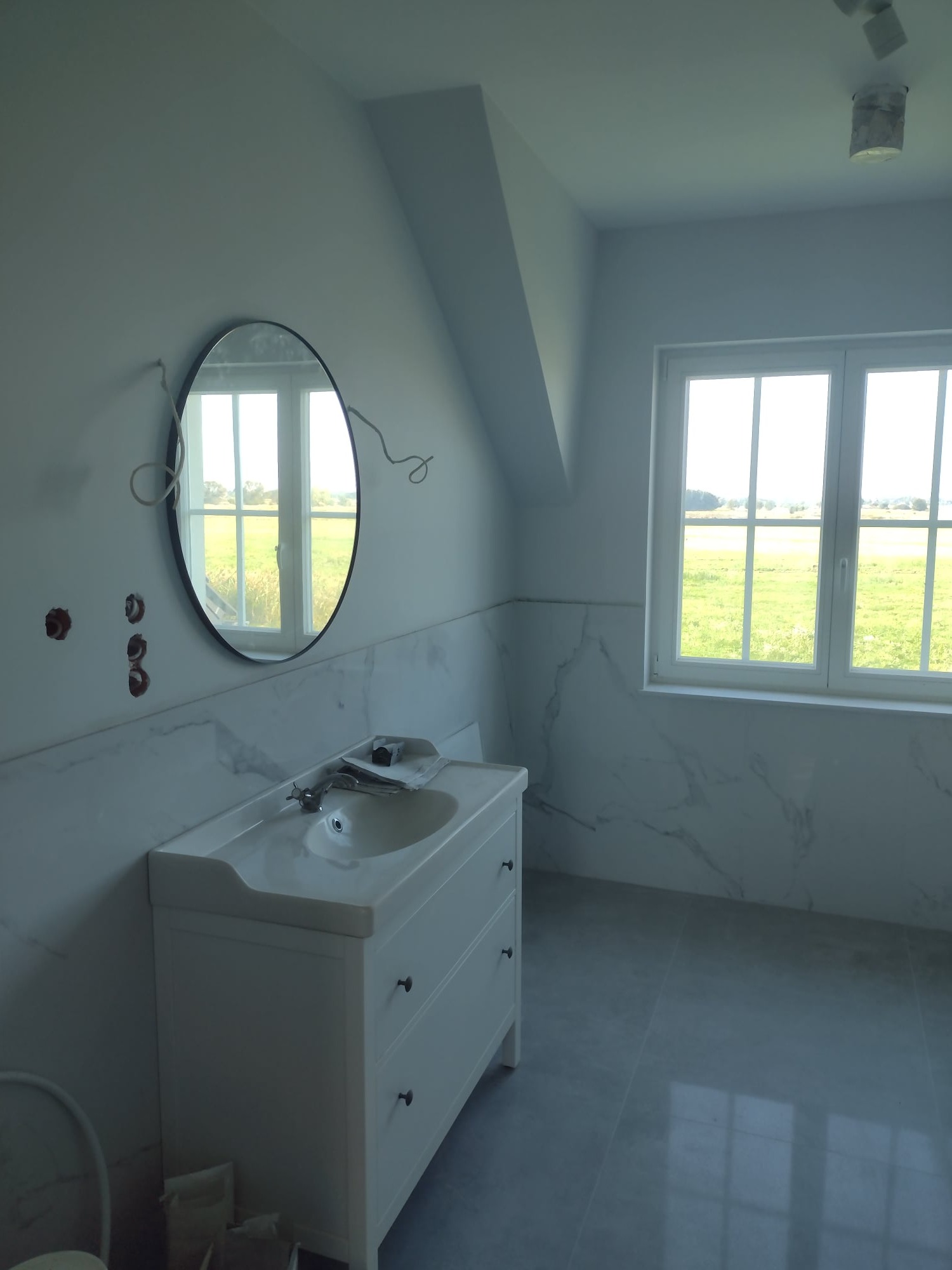 Nowoczesna łazienka w trakcie wykańczania: biała szafka z umywalką, lustro w czarnej ramie, marmurowe płytki na ścianach oraz wystające kable ze ściany.