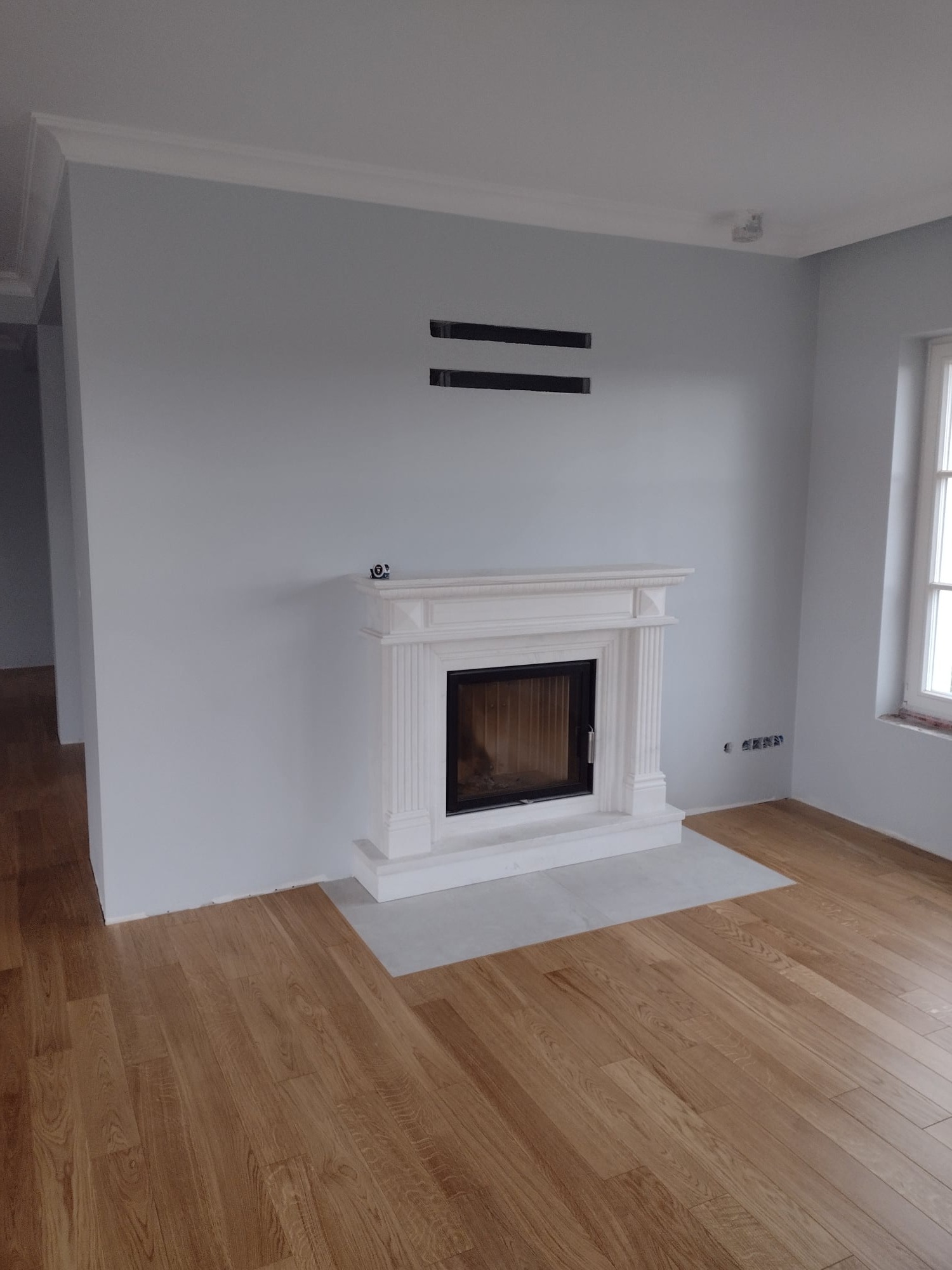 Jasne wnętrze z drewnianą podłogą, marmurowym kominkiem z czarnym wkładem, szare ściany z listwami przysufitowymi i niezamaskowanymi otworami wentylacyjnymi.