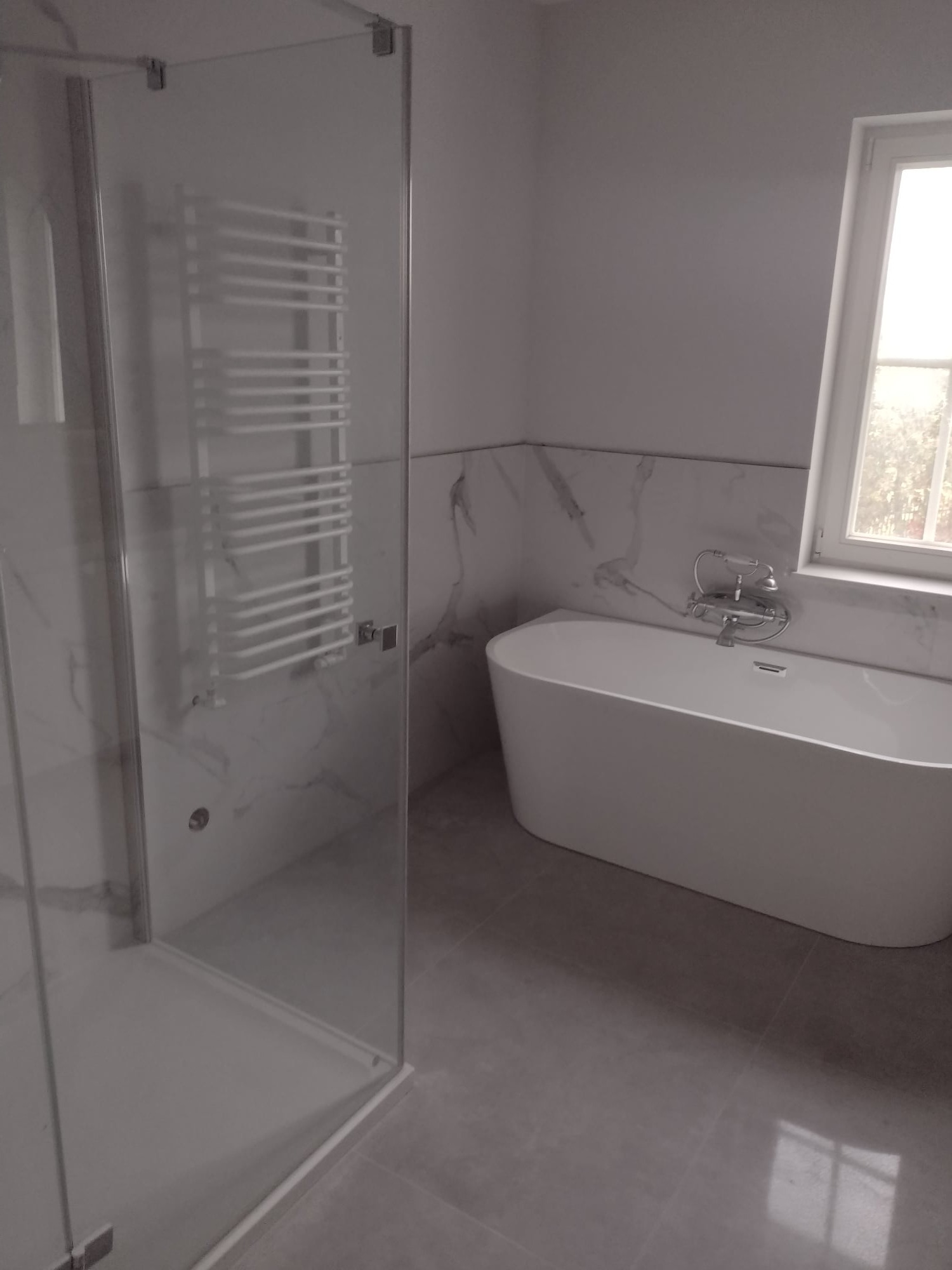 Nowoczesna łazienka z wolnostojącą wanną, szklaną kabiną prysznicową z grzejnikiem drabinkowym i marmurowymi płytkami ściennymi.