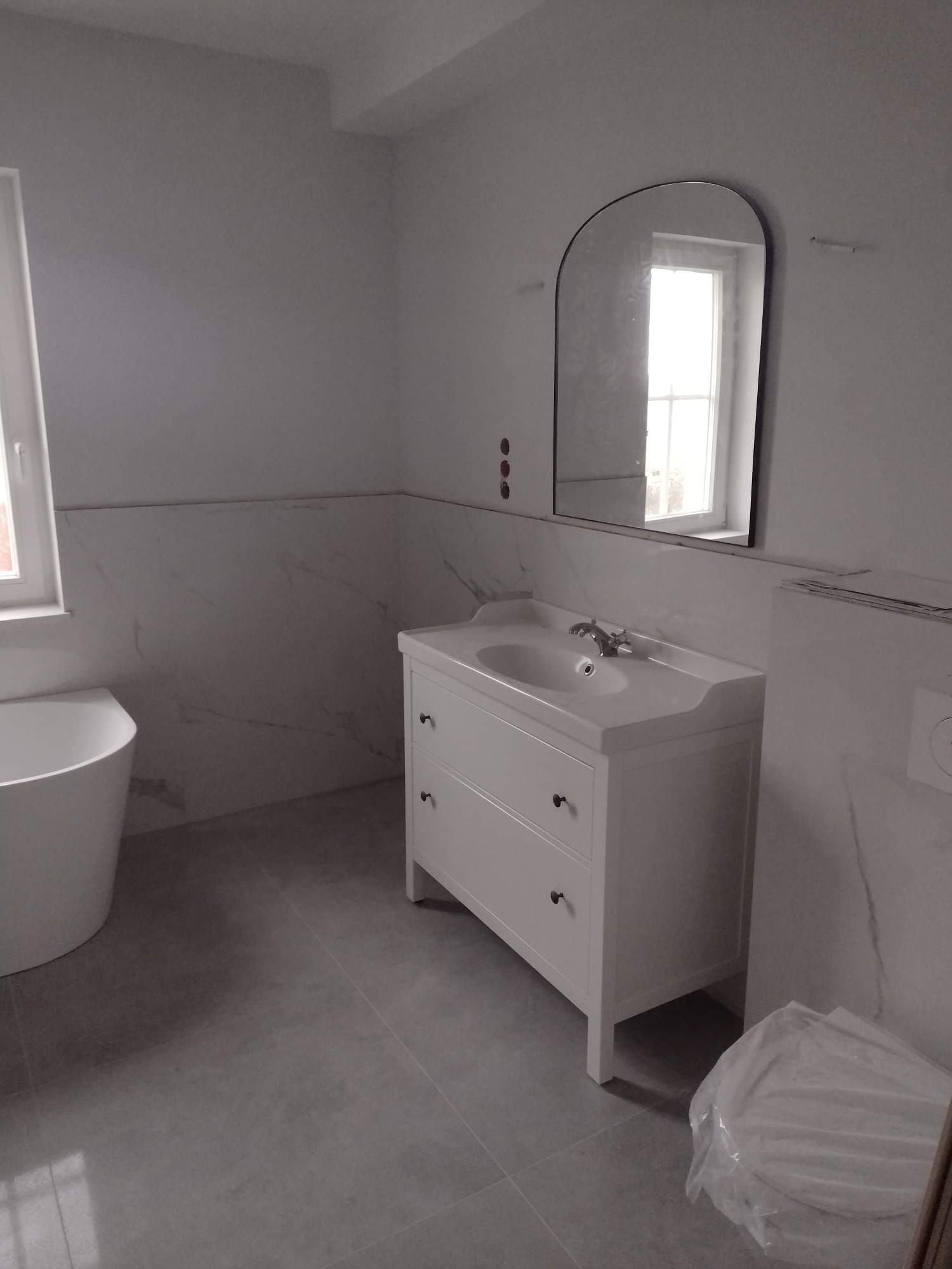 Biała łazienka w trakcie wykańczania: owalna wanna, szafka z umywalką i owalne lustro, płytki imitujące marmur na ścianach, szare płytki podłogowe, widoczne ślady po montażu lustra, brakujące...