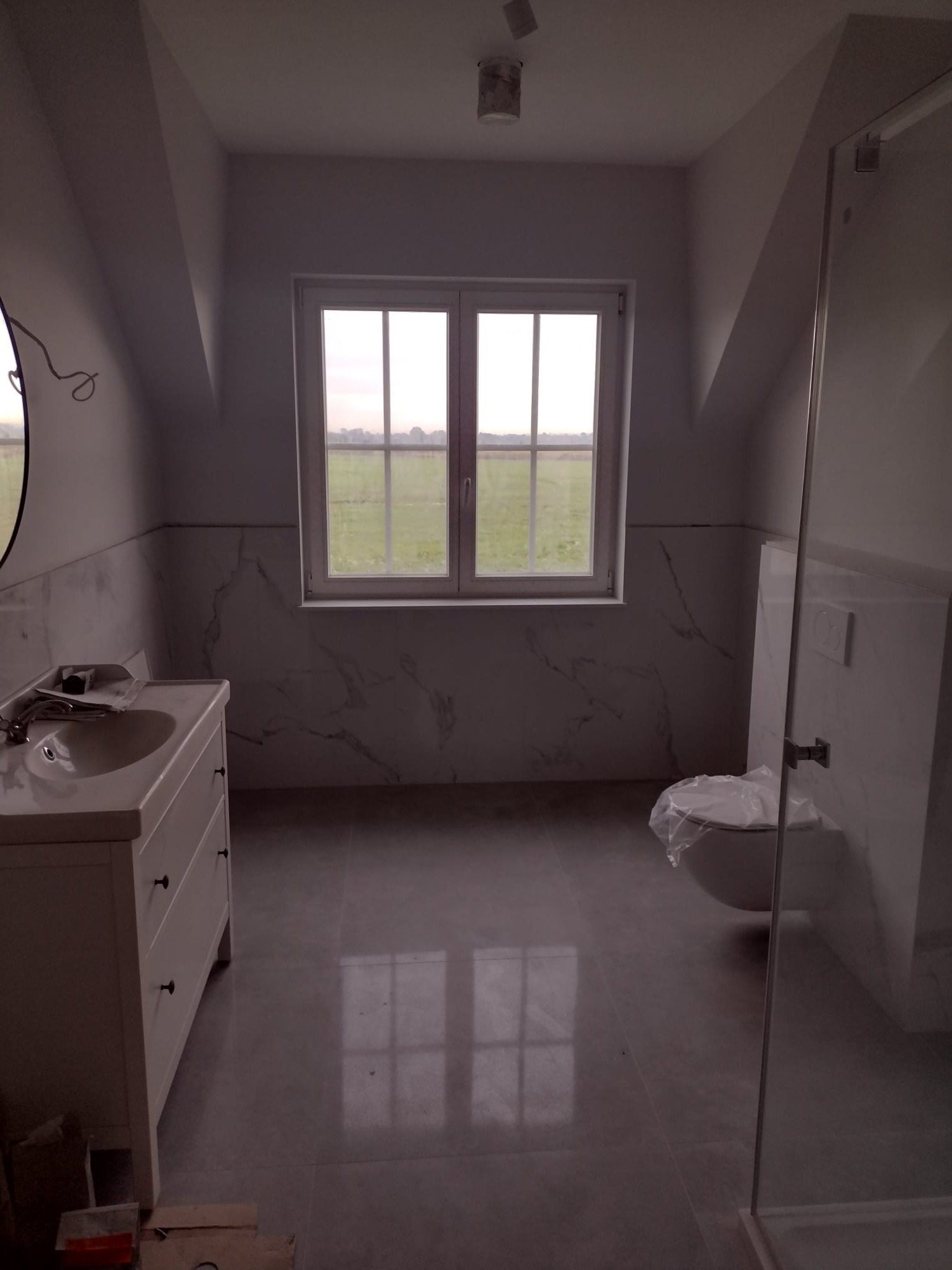 Jasna łazienka na poddaszu z oknem na krajobraz, marmurowe płytki na ścianach, biała szafka z umywalką i toaleta z folią ochronną, szklana kabina prysznicowa.