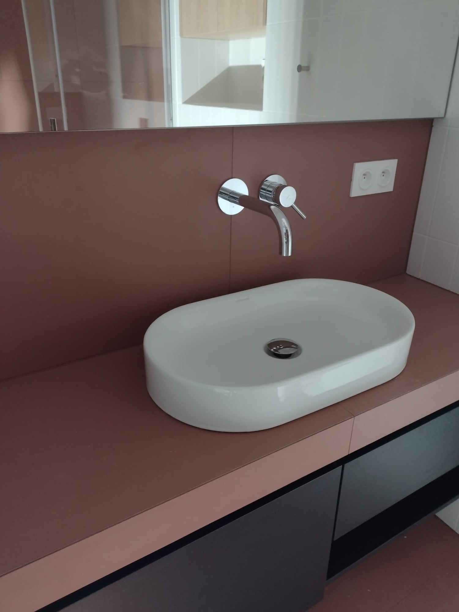 Nowoczesna umywalka nablatowa o owalnym kształcie i chromowana bateria na tle ściany w kolorze terakoty z widocznymi gniazdkami elektrycznymi oraz fragmentem lustra i kabiny prysznicowej w tle.