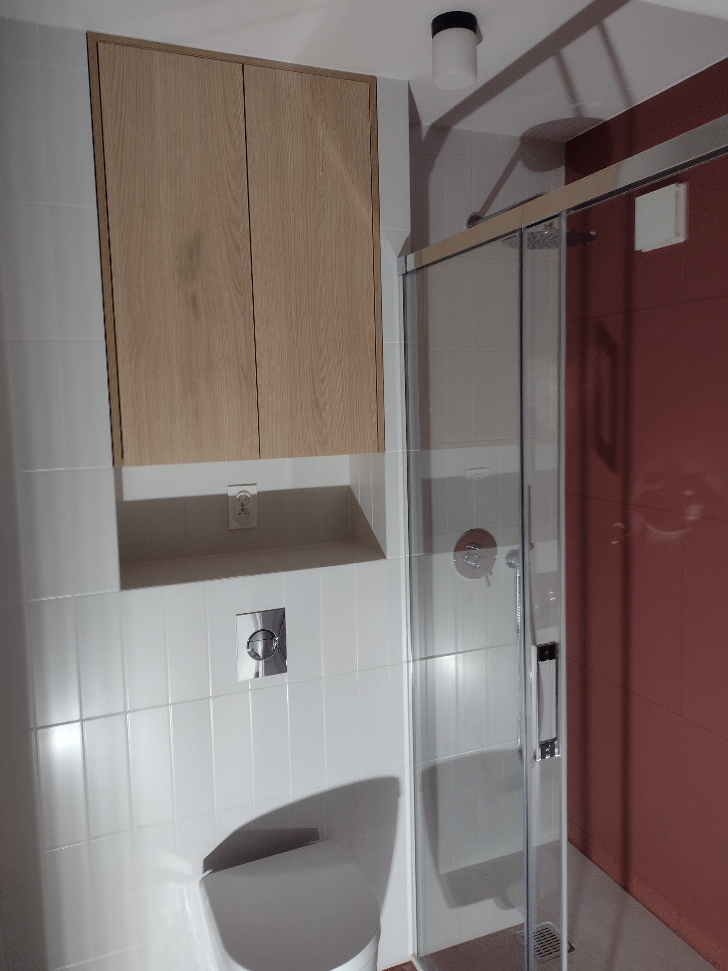 Nowoczesna łazienka po remoncie: białe płytki, zabudowa z drewnianymi szafkami nad toaletą, kabina prysznicowa z przesuwnymi drzwiami i czerwona ściana wewnątrz.