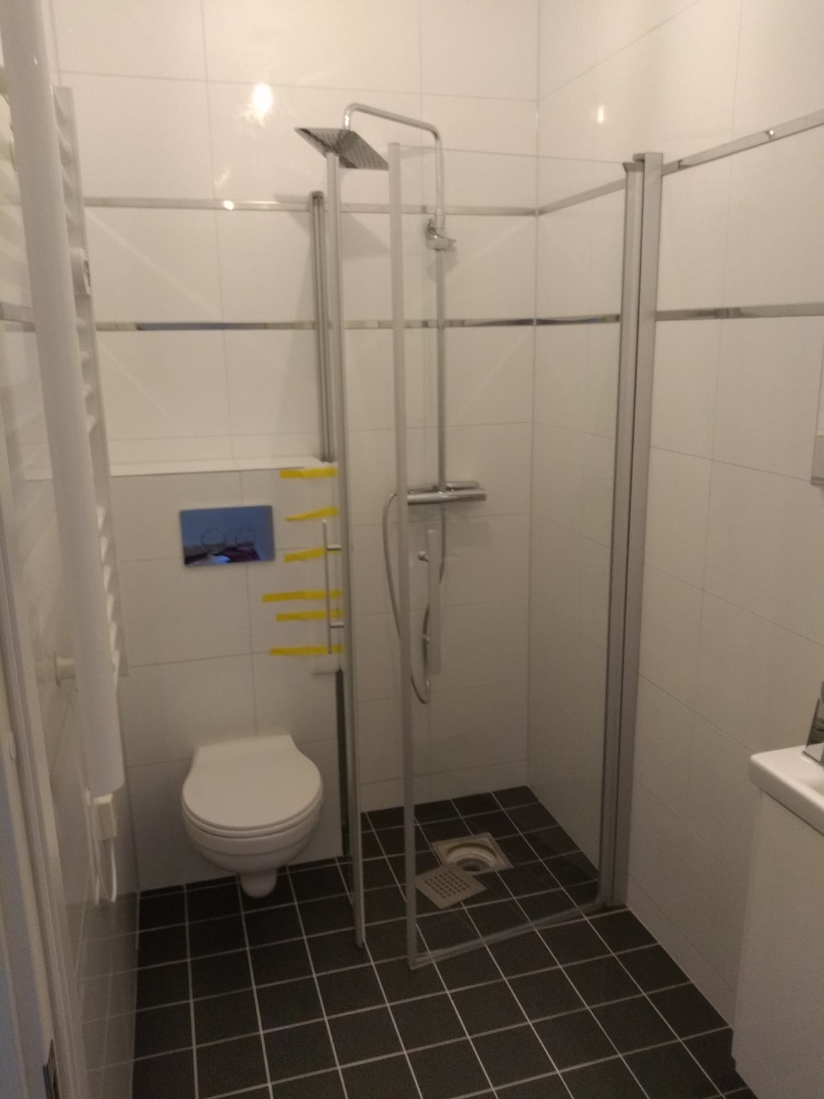 Nowoczesna kabina prysznicowa z deszczownicą, białe płytki ścienne z metalową listwą, ciemna podłoga z kwadratowych płytek, wisząca toaleta z niebieskim przyciskiem spłukującym, żółte taśmy...