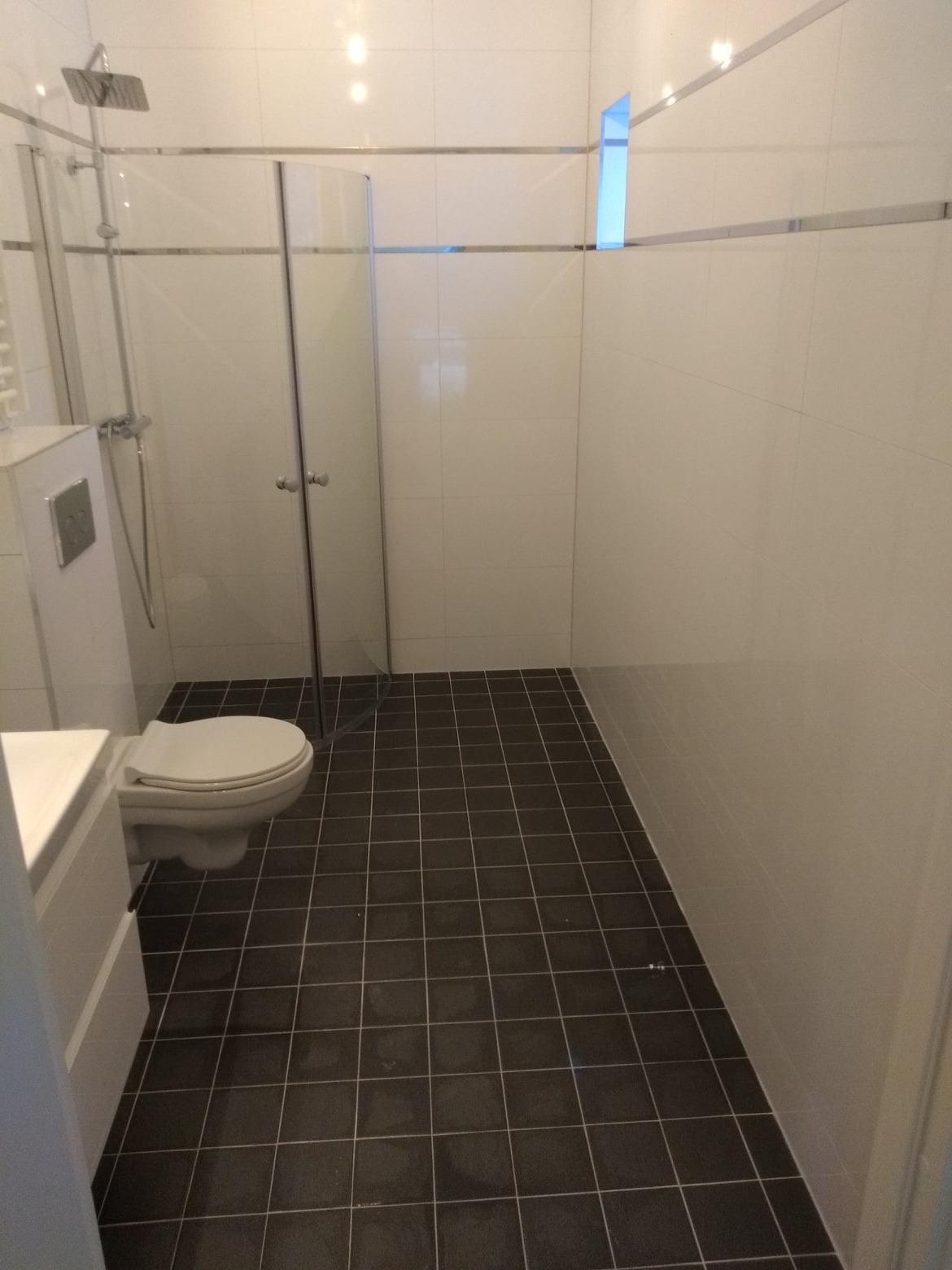 Nowoczesna łazienka z kabiną prysznicową typu walk-in, białymi płytkami ściennymi z metalicznymi listwami i ciemną, kwadratową terakotą na podłodze, widok na toaletę i umywalkę.