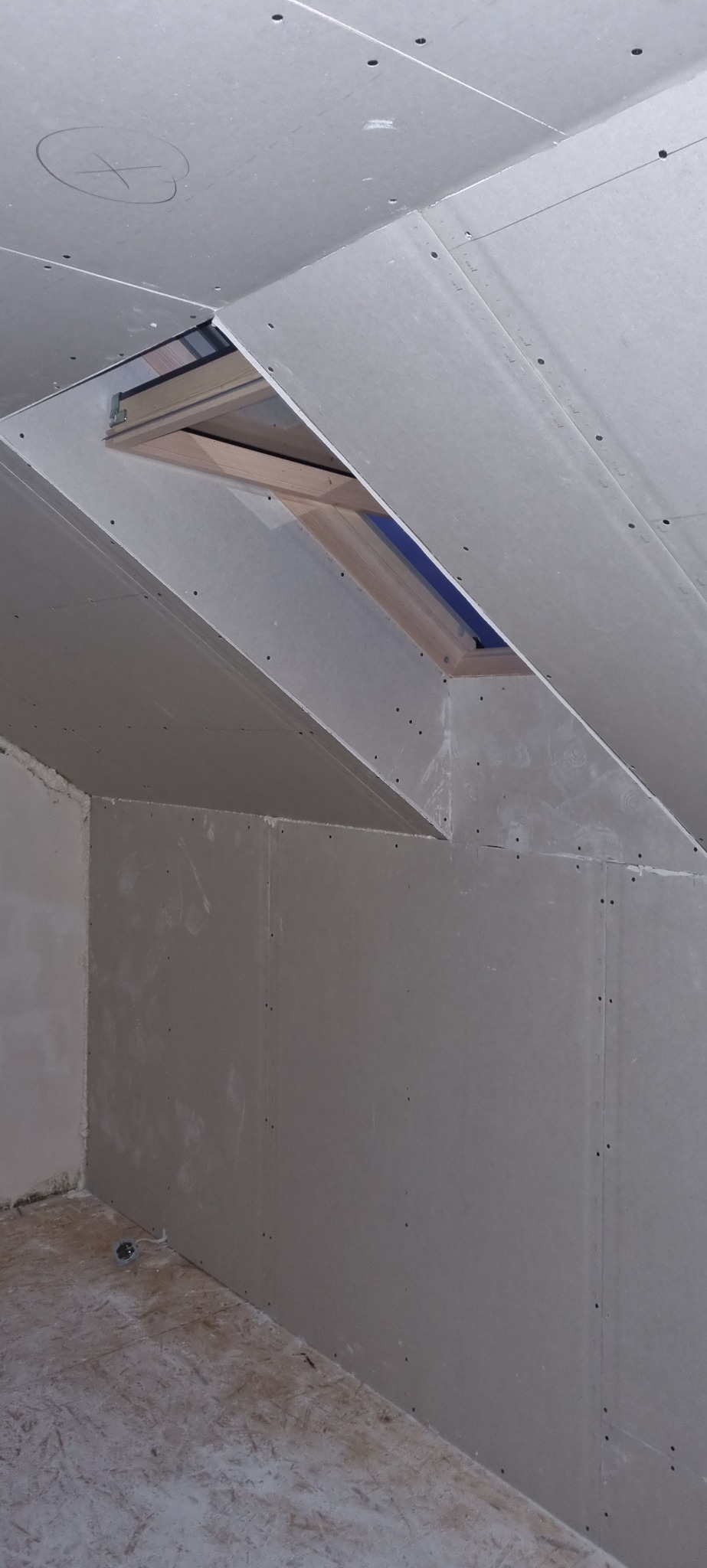 Montaż płyt gipsowo-kartonowych na poddaszu z oknem dachowym, widoczne łączenia płyt i ślady szpachlowania, przygotowanie do dalszych prac wykończeniowych.