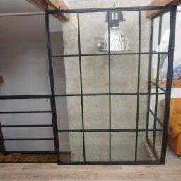 PROFIL - Nowoczesna balustrada schodowa z czarnych metalowych profili i szkła, oddzielająca przestrzeń schodów od salonu z żółtą sofą, widoczna lampa w stylu industrialnym i drewniane elementy konstrukcyjne.