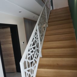 PROFIL - Klatka schodowa z drewnianymi stopniami i białą, ażurową balustradą z geometrycznym wzorem, widziana z dołu, z lampą ścienną w tle.