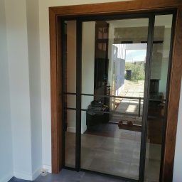 PROFIL - Przeszklone drzwi przesuwne z czarną metalową ramą i drewnianą ościeżnicą, widok na taras i ogród.