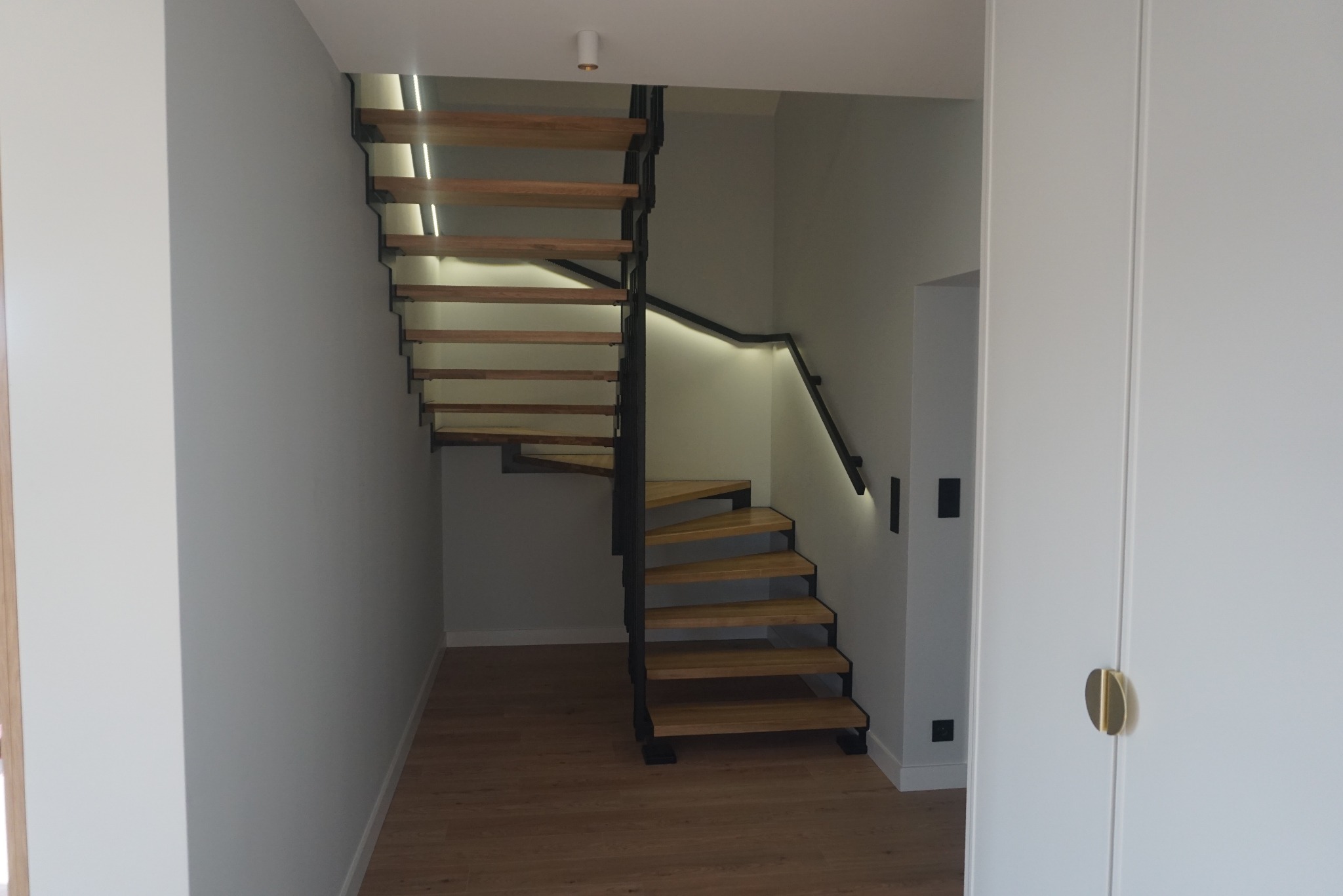 Nowoczesne schody z drewnianymi stopniami i czarną metalową konstrukcją, podświetlone LED, w minimalistycznym wnętrzu z białymi ścianami i drzwiami.
