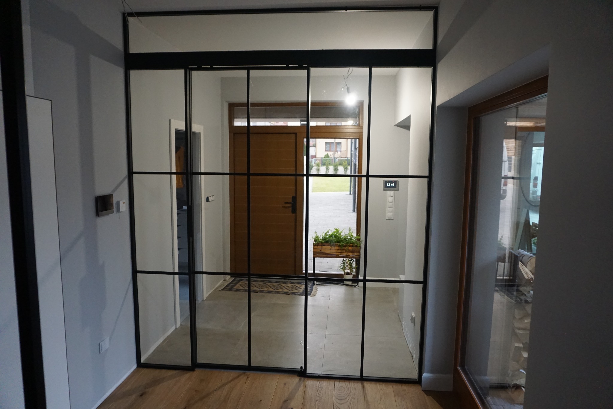 Przeszklona ściana działowa z czarnymi profilami, tworząca efekt loftowy w nowoczesnym wnętrzu. Widoczny fragment korytarza z drewnianymi drzwiami wejściowymi i roślinami w donicach.