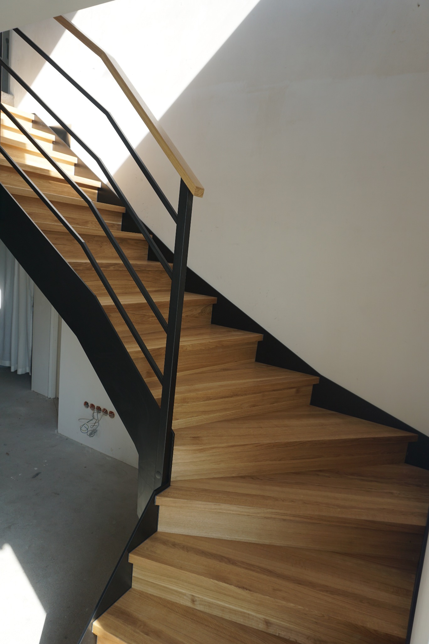 Drewniane schody z czarną metalową konstrukcją i jasną drewnianą poręczą, oświetlone naturalnym światłem w trakcie montażu, widoczne puszki elektryczne w ścianie.