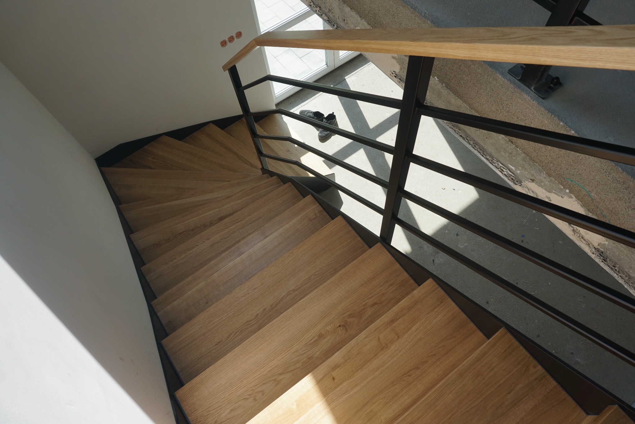Spiralne schody z drewnianymi stopniami i czarną metalową barierką z drewnianym poręczem, widok z góry, na podeście leżą buty.