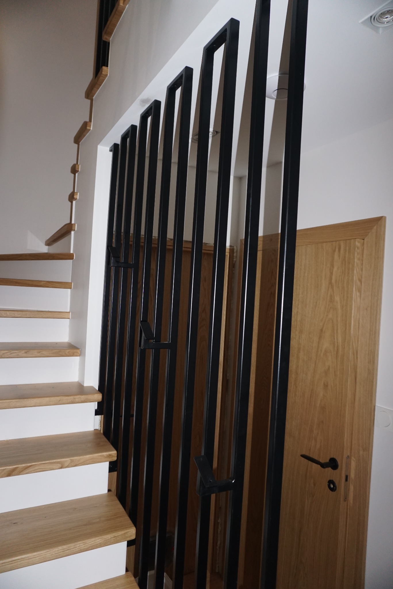 Nowoczesne schody drewniane z białymi podstopnicami i czarną, minimalistyczną balustradą metalową z pionowych profili, widok z dołu, drzwi drewniane w tle.