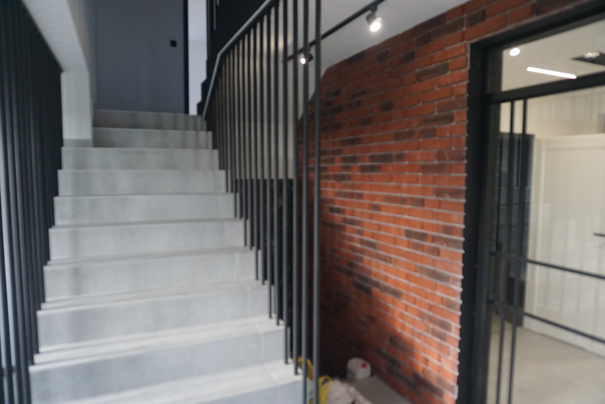 Betonowe schody wewnętrzne z czarną, pionową balustradą w Suwałkach, na tle ceglanej ściany i metalowej konstrukcji bramy.