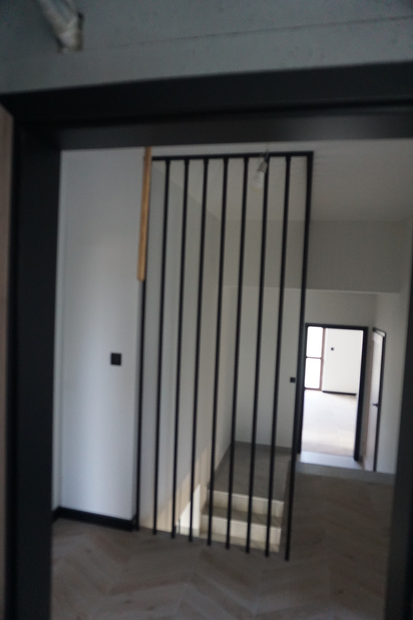 Nowoczesne schody z minimalistyczną, czarną balustradą z pionowych prętów, widoczne zza czarnej ramy drzwiowej, z perspektywą na dalsze pomieszczenia.