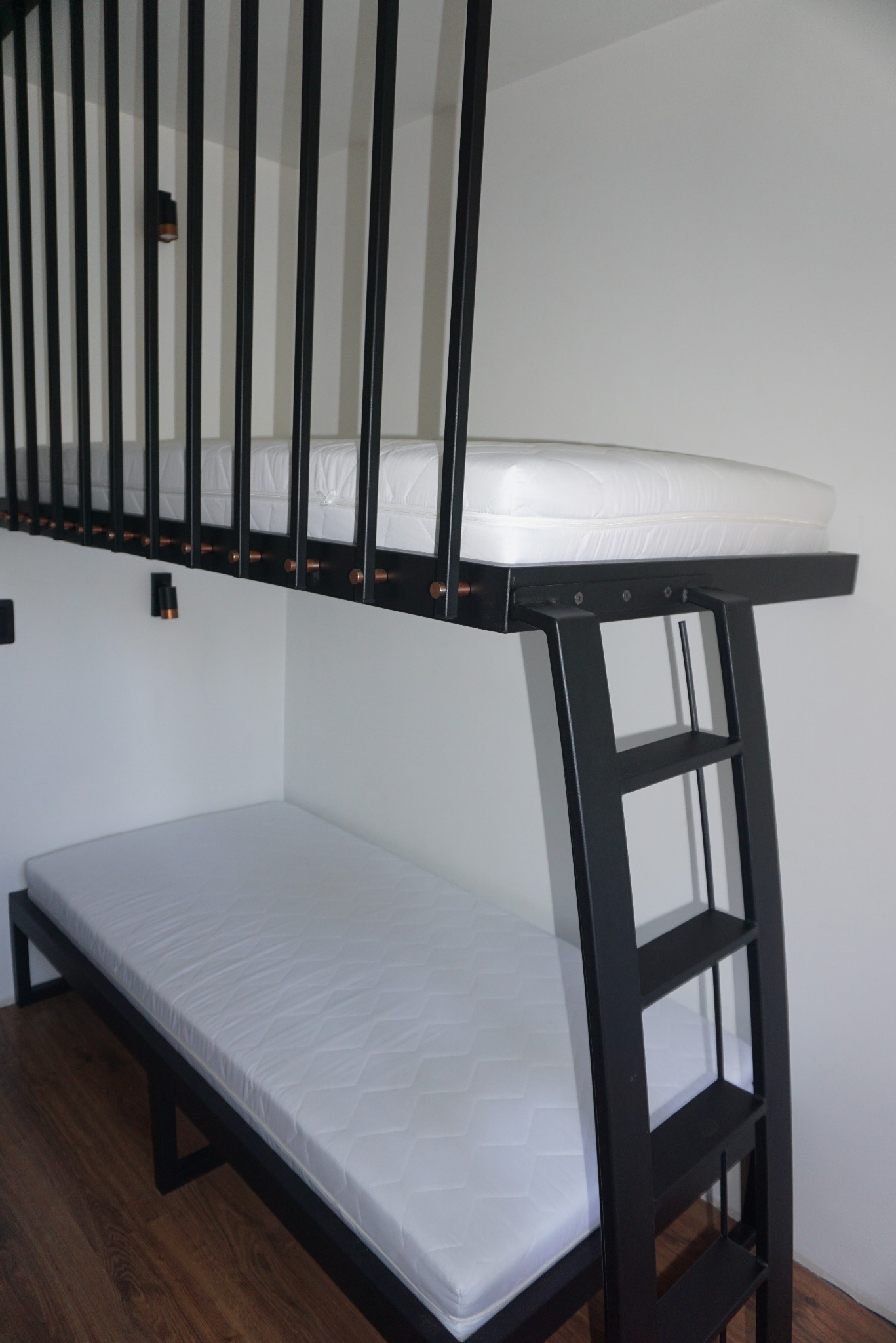 Dwuosobowe łóżko piętrowe z czarną metalową ramą i drabinką, górna część zabezpieczona pionowymi prętami, minimalistyczny design, materace w kolorze złamanej bieli.